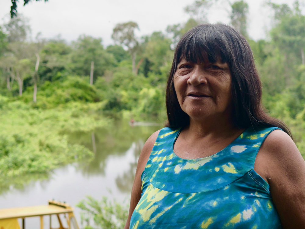 An indigenous woman, who looks over 50 years old, with straight hair and bangs, wearing a blue and yellow dress. She is looking at the camera.