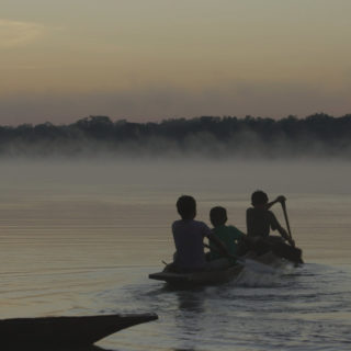 Imagen de la silueta de tres niños en una canoa, remando en un río, en dirección a la selva. La foto parece haber sido tomada al anochecer.
