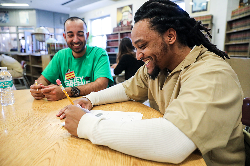 PHOTO 8 - In the foreground, in profile, is a black man with dreadlocks and a goatee, who appears to be between 30 and 40 years old, wearing a long-sleeved white shirt under a short-sleeved beige shirt. He is taking down notes on a piece of paper and smiling. In the background, which is out of focus, a slightly balding, young white man wearing a green T-shirt also smiles, looking at the sheet of paper.