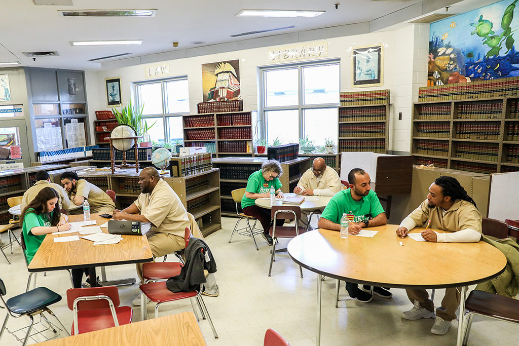 A room (study room or library), with tables, some rectangular and some round. Some people are wearing green shirts (volunteers) while others wear uniforms with beige shirts and pants (prisoners). Most are taking notes and talking. In the background are several bookshelves with books (including encyclopedias) and globes. There are colorful pictures on the walls.