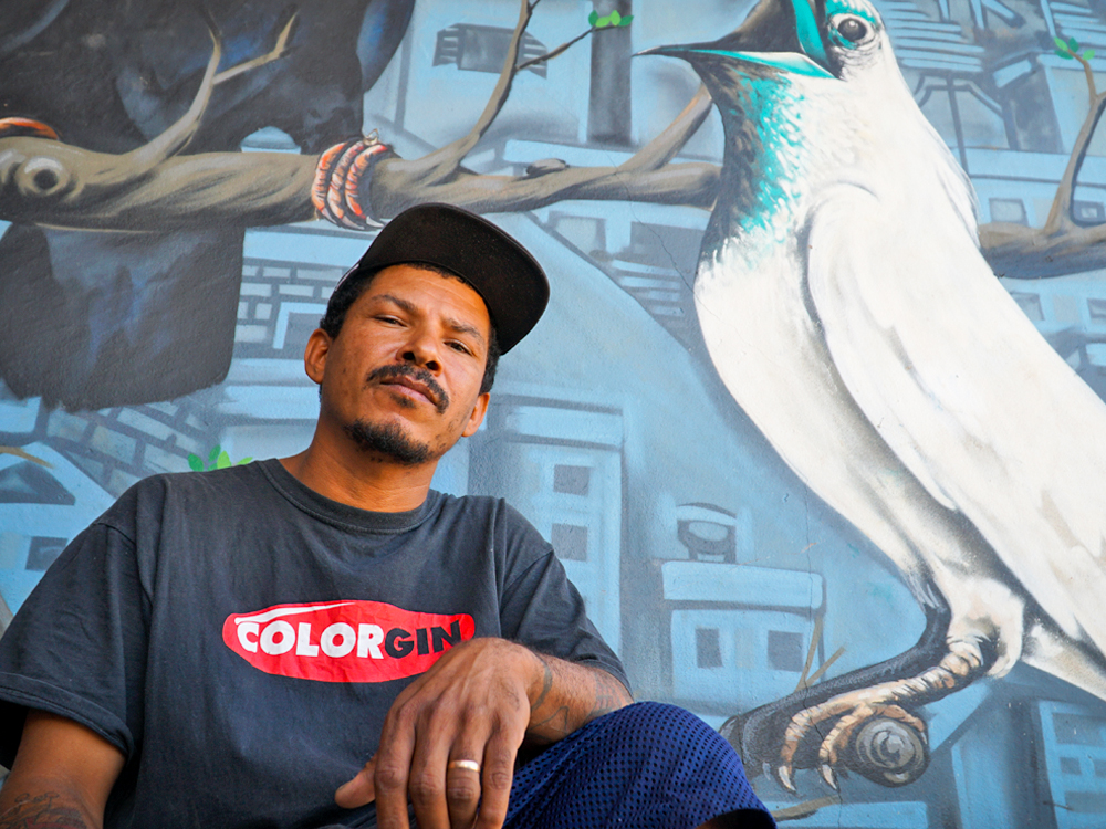 A black man with a beard and mustache is wearing a black cap and a black T-shirt. He is looking at the camera, with a serious expression. Behind him, on a wall, is a graffiti mural depicting a large bird on a tree branch, with apartment buildings in the background.