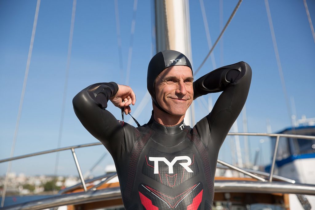 A middle-aged, skinny white man is smiling at the camera. He is wearing a tight black swimming suit with long sleeves and the letters "TYR" printed in white on the chest, as well as a black swimming cap with a gray stripe across, with the same letters printed on it. The man has his arms back over his shoulders, trying thin black strings over his back. In the back is the mast and front of the boat on which he is standing, and a bright blue sky.