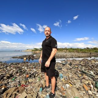 A thin man with white skin, short gay hair and beard, wearing all black (polo shirt, shorts and sneaker) is standing facing the left side of the photo and looking at the camera. The ground around him is covered in plastic waste, ending in the sea behind him. At the top of the picture, above the landscape, is a bright blue sky.