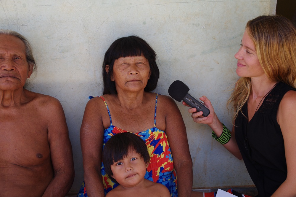An elderly, indigenous woman with black and straight hair and bangs, she is wearing a colorful dress and looking at the camera. On her lap, there is indigenous boy without shirt. On the left side of the picture, an old man, with white hair and no shirt. Both are sat, their backs are against the white wall, and they're looking at the camera. On the right side of the image, there is a white woman, with long blond and wavy hair, she is sitting sideways to the camera and looking at the couple of natives. She holds a microphone with her right hand.