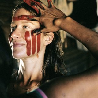 A white woman, with blond and wavy hair and light eyes is looking at the right corner of the image. She has her left cheek painted with three red stripes. In the foreground, in the left corner of the image, an indigenous woman appears on her back. You can only identify part of her hair and left arm with her fingers in red and black paint. She's painting the white woman's face.