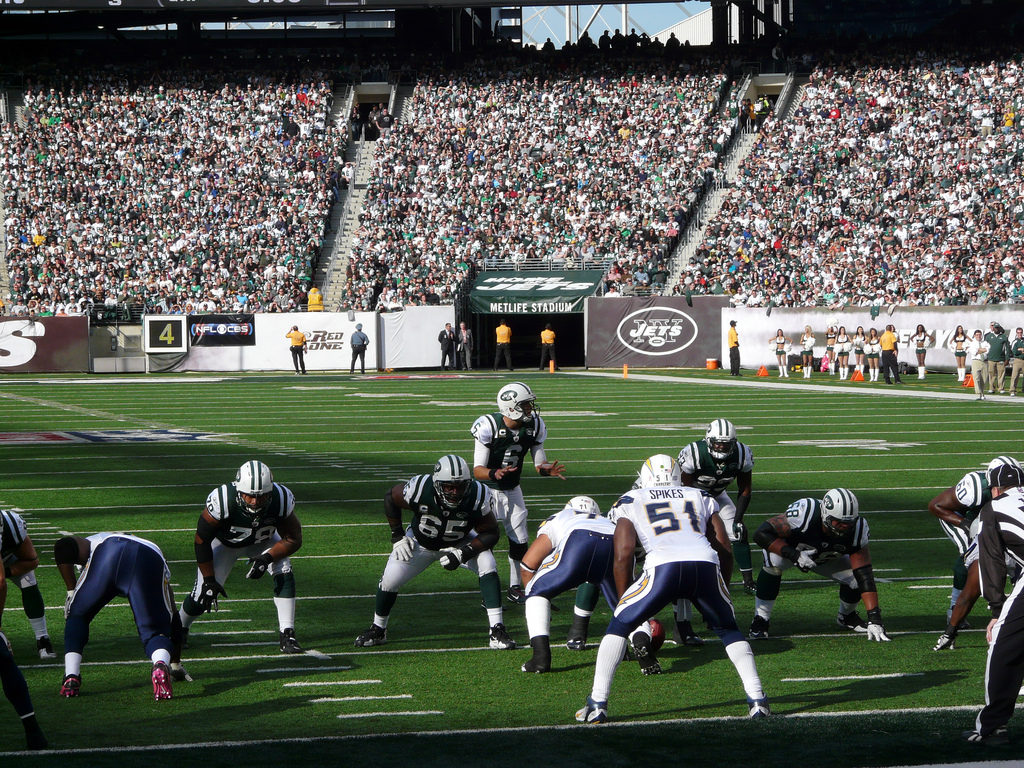 Ten american football players, wearing uniform consisting of tights, short sleeve shirts and helmets, are lined up for a play inside a sports stadium. The field is covered in low green grass. The uniform of the players with their backs to the camera is made of dark blue pants and white shirts, while the other team's is white pants and green shirts. All of their helmets are white. In the back right corner, cheerleaders in white are gathered. Further back are the stands filled with people. The area is sunny.
