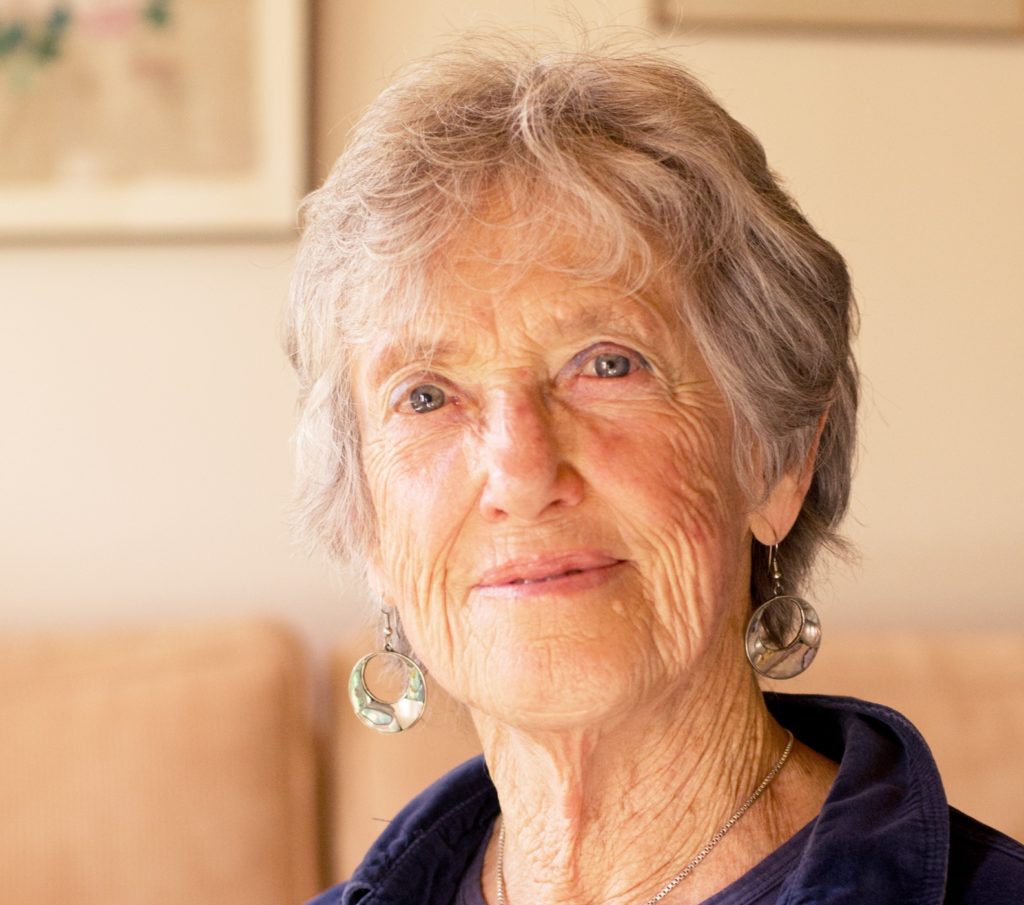 Uma mulher idosa, cabelos grisalhos e curtos, olhos claros, usando uma camisa azul e brincos de pequenas argolas metálicas, olha fixamente para a câmera. A imagem apenas mostra dos seus ombros para cima. Ao fundo, imagem desfocada de uma sala de estar.
