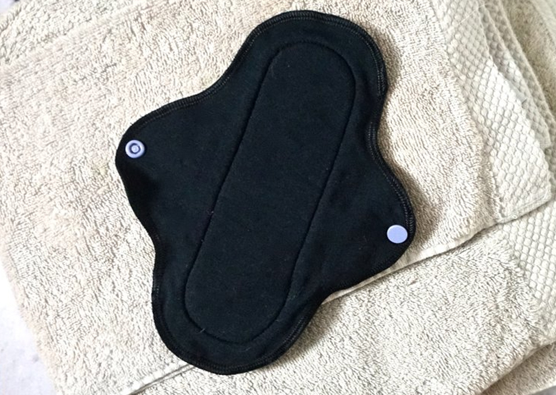 A feminine hygiene pad made from black cotton with small light blue buttons is open on a white towel that is folded into a square.