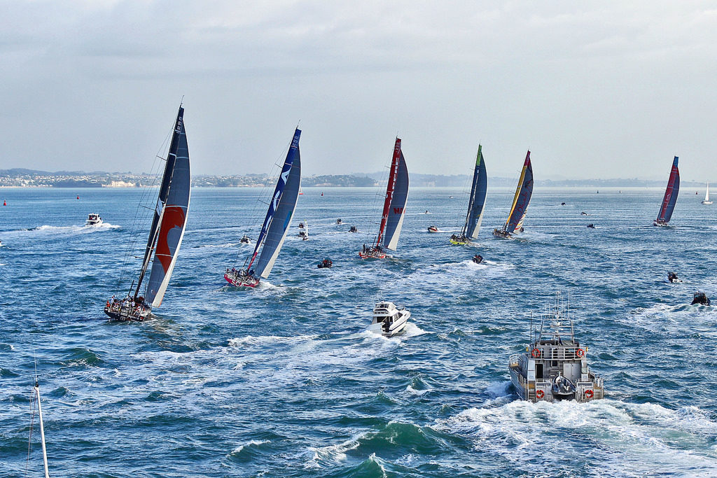 Six small sailboats are on a clear blue sea, with large white sails with print on them. Around the boats are some small motorboats. In the background is a slightly cloudy sky.