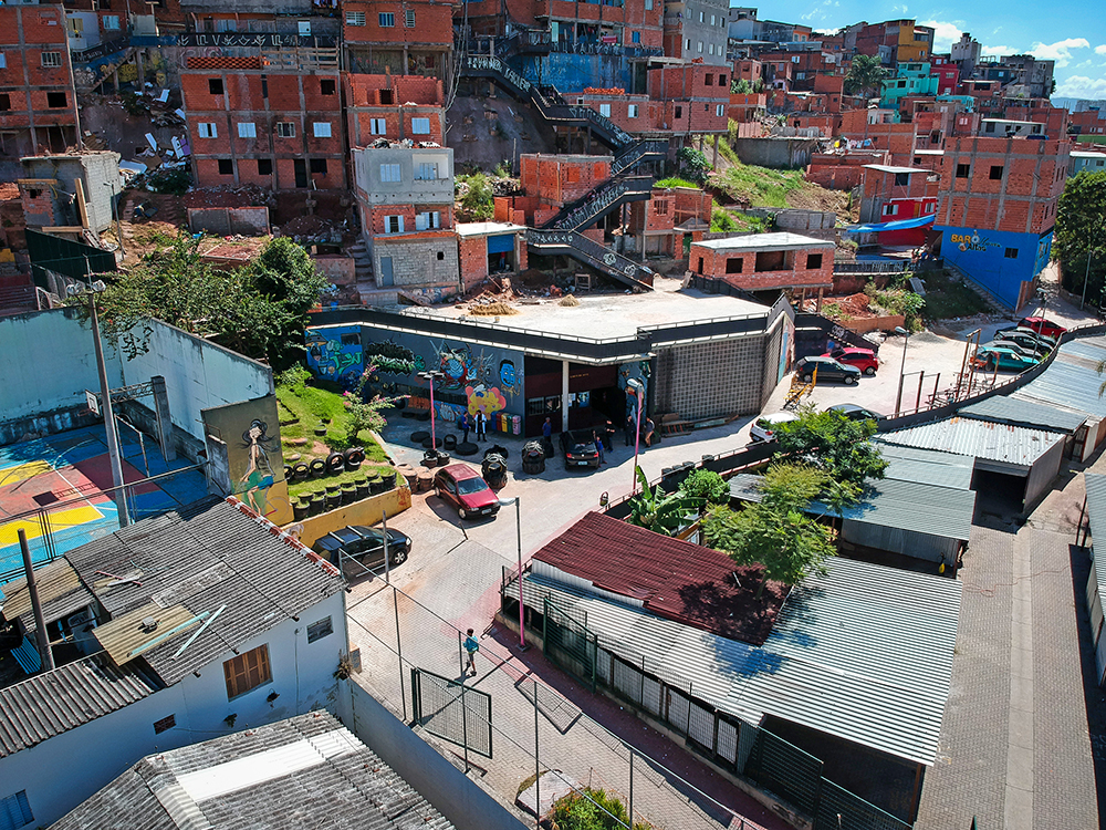 An aerial image of a dense suburban neighborhood, with small brick houses in full view on a hill. In the center of the photo is a small shed with graffiti on its front outside wall. In the foreground is a quiet street with some parked cars.