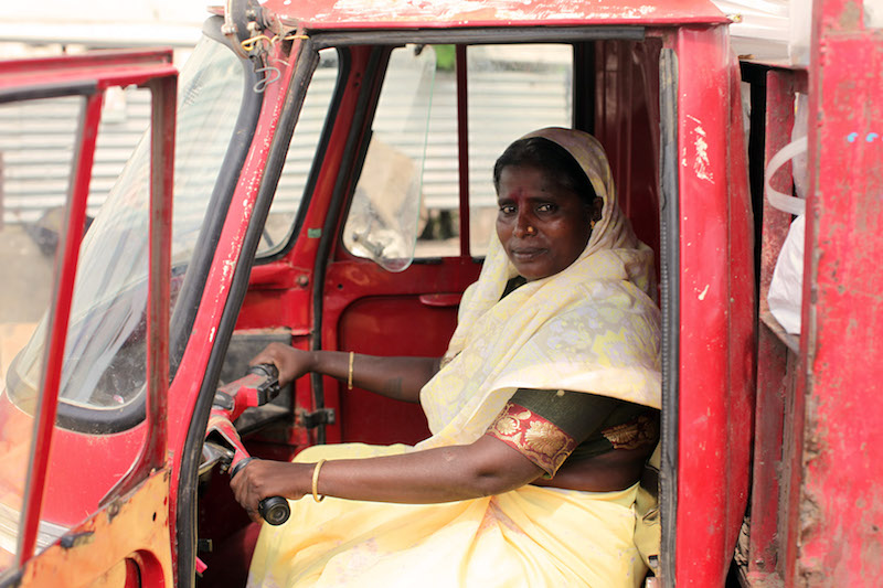 Una mujer de piel negra, ojos oscuros y pelo liso oscuro, viste un sari (vestido fino) traslúcido de color arena que le cubre la cabeza y los hombros, además de las piernas a modo de falda. Lleva también una camiseta verde oscuro metalizado, con una franja roja con estampado dorado en la parte final de la manga, encima del codo, y finas pulseras doradas. La mujer está en el asiento del conductor de una camioneta roja, con cada mano en el manillar del vehículo, y el cuerpo de frente hacia el lateral izquierdo de la foto, mientras mira de manera expresiva a la cámara, con la cabeza girada. El camión tiene la puerta izquierda, que está más cerca de la cámara, abierta. Al fondo, la puerta derecha del camión, cerrada.