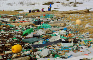 La foto muestra una playa cubierta con una variedad de desechos de plástico. En la parte de atrás hay tres personas que llevan abrigos pesados con el suelo cubierto de nieve.
