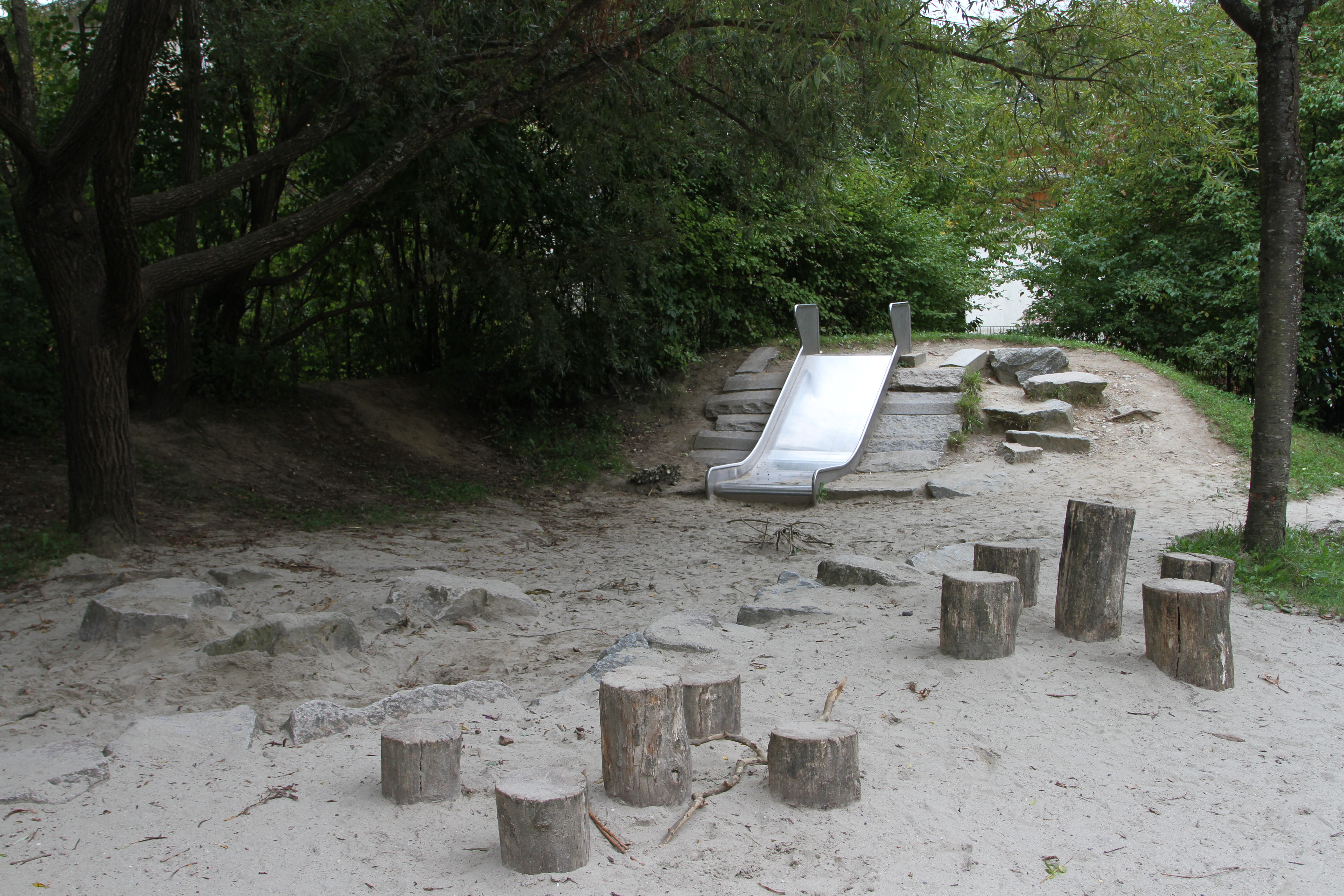 Photo of a metal slide attached to the side of a small hill. Tree stumps in front of it serve as benches. Surrounding this small clearing, green, leafy trees.
