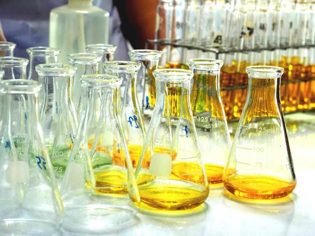 Imagen de un laboratorio con varios frascos transparentes. Los que están a la izquierda están vacíos, en el medio y a la derecha tienen una pequeña cantidad de líquido amarillo y otros verde. Al fondo y a la derecha, tubos de ensayo llenos del mismo líquido amarillo.