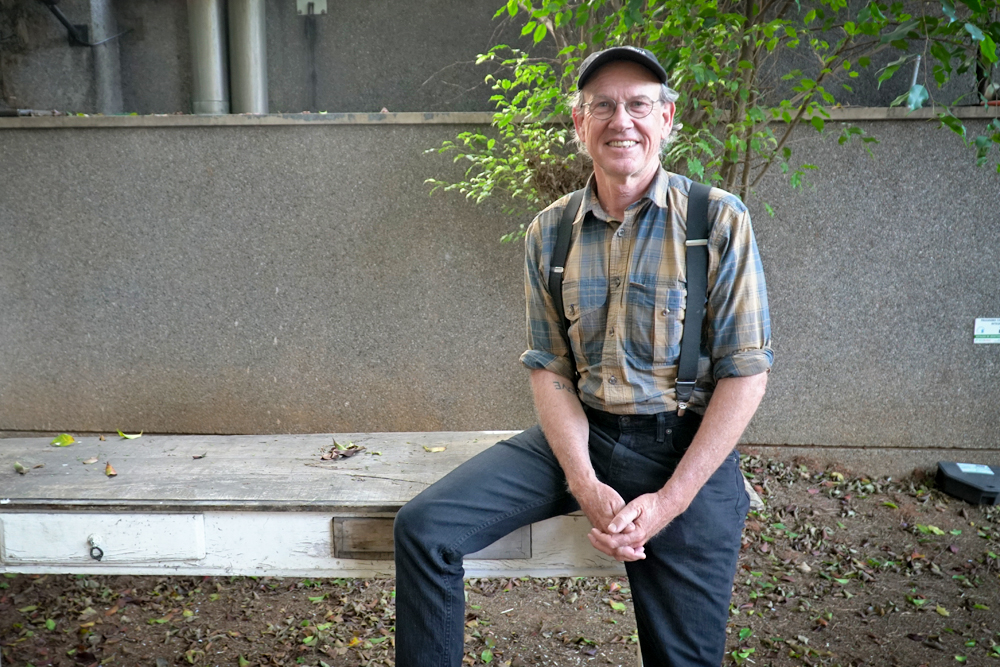 A white man with white hair, wearing a cap and glasses, looks at the camera and smiles. His hands are clasped together. He is standing, but resting his right leg on a wooden bench. He wears a yellow and blue checked shirt with black suspenders and dark blue pants. Behind him is a cluster of small leafy trees with thin trunks, a gray wall and some leaves that have fallen from the tree onto the ground.