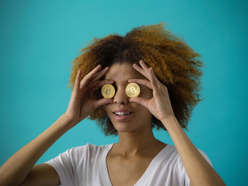 A thin black woman wearing a white t-shirt is holding up two coins with a B (the symbol for Bitcoin) over her eyes. Her hair is curly and blonde. The background is blue.