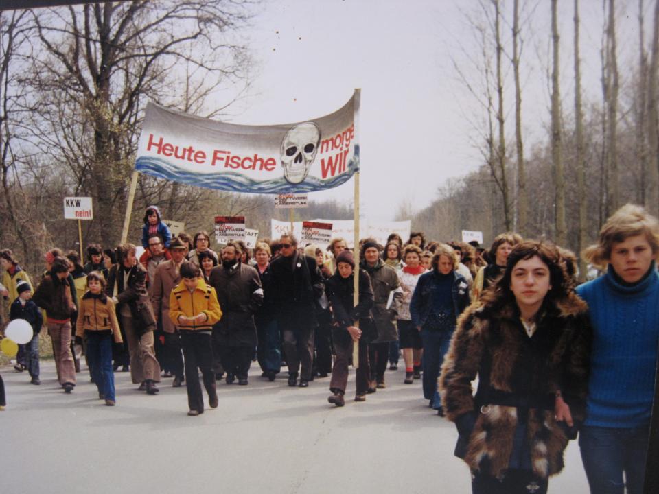 Um grupo de pessoas vestidas com roupas de inverno está caminhando. Eles seguram uma faixa com a inscrição, em alemão "Hoje peixe, nós amanhã" e o desenho de uma caveira ao lado do "nós amanhã".