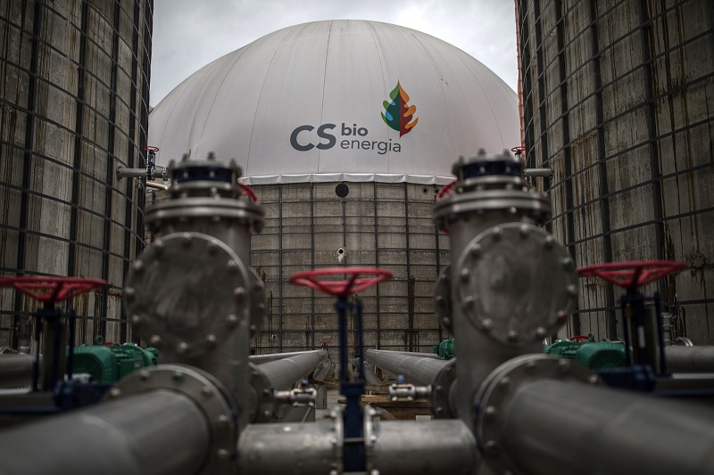 At the center of the photo, towards the back and in focus, is the white dome of a chemical plant. Written in black is the name "CS bioenergia," along with the company's logo - a blue droplet below a pine tree that is half green, half orange. There are two tall gray walls flanking the dome on each side, and metal piping in the foreground.