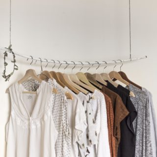 Against a white background, thirteen wooden hangers with women's clothing rest on a white metal rod suspended from the ceiling by two pieces of steel wire.
