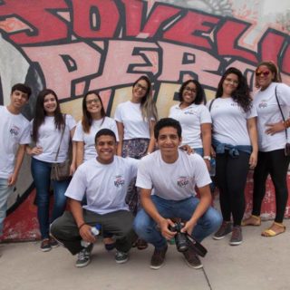 Nine youngsters smile at the camera while posing against a colorful mural, all wearing a white t-shirt with the "Fala Roça" logo printed on the left side. Two young men are crouching in front. The one of the left has his arm around the one on the right, who is holding a camera. Behind them is a young man on the left and six girls next to him, all in a row