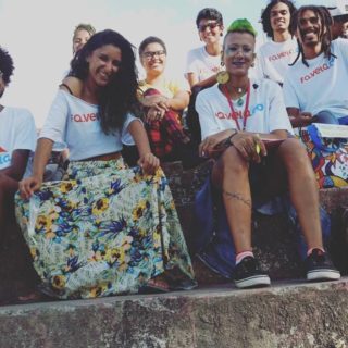 Um grupo de nove jovens, integrantes do FA.VELA, usam camisetas com o logo do grupo. Eles estão sentados em uma escada e sorriem para a foto.