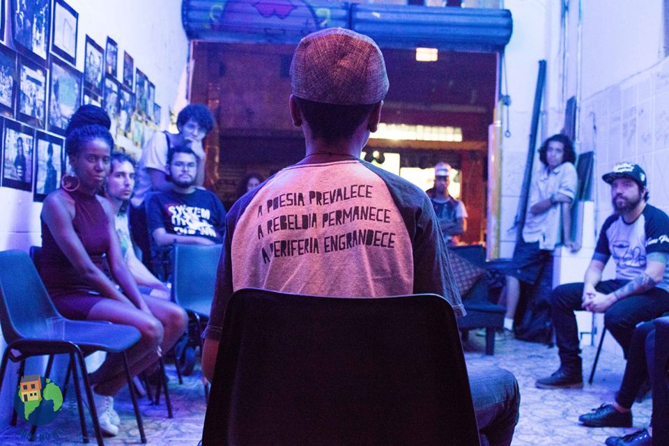 On the foreground is a young black man, sitting with his back to the camera, wearing a t-shirt with the phrase "Poetry prevails. Rebelliousness remains. The periphery grows." written in Portuguese. He is wearing a pink cap and is facing a circle of people in a space lit by a blue light and with many photographs on the wall. The people are all looking at the young man.