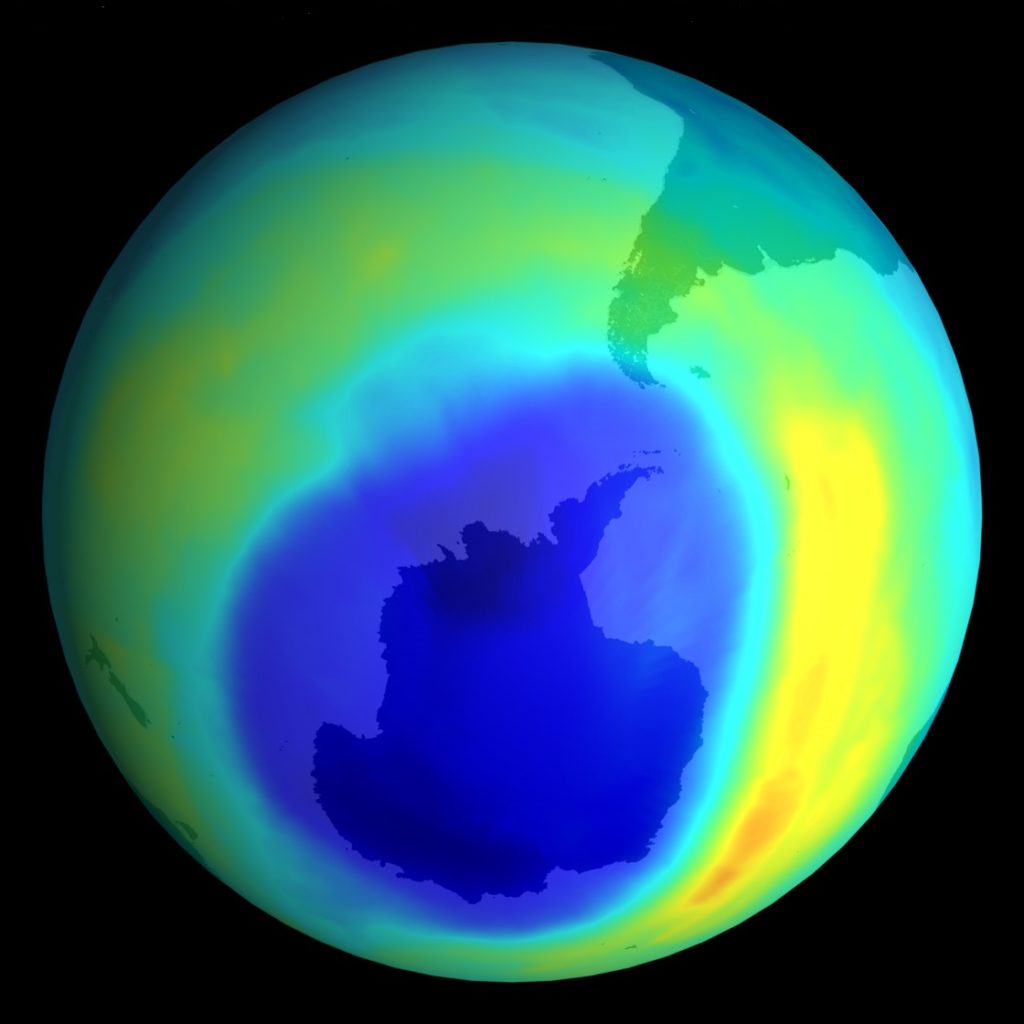 Planeta Terra, representada por um globo em diferentes cores brilhantes - amarelo, verde, azul claro e azul escuro. Uma bolha azul mais escura e disforme aparece quase no centro da imagem, representando o buraco na camada de ozônio sobre a Antártida.
