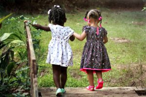 Two small, black girls are holding hands, their backs to the camera. They're crossing a small, wooden bridge onto a green lawn. The darker-skinned girl, who is on the left side, has her hair tied into a ponytail, with a white ribbon on top. She wears a white dress with a colorful pattern, as well as pale green shoes. Her left arm is outstretched. Her companion, to her right, is a lighter-skinned black child whose hair is divided into braids, a hot pink ribbon-shaped barrette on the end of each one. She wears a black dress with a pink and white pattern, and pink trim. Her shoes are also pink. The pink in the barrette, dress and shoes is the same shade.