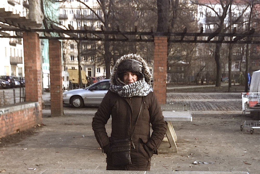 A white-skinned woman is wearing a heavy brown winter coat, a black and white scarf and a hat. She is in the center of the photo and is looking at the camera. The image shows her from the knees up. In the background is an urban area: brick columns, a parked car and some trees.