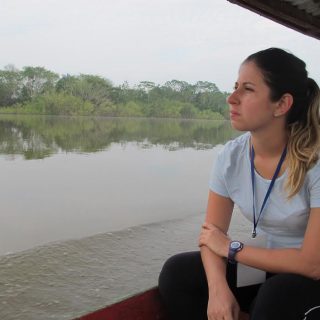 Uma mulher jovem, aparentando 20 e poucos anos, loira de cabelos presos, veste uma camiseta cinza e uma calça preta. Ela está sentada na beira de um barco, observando o rio e a paisagem verde ao redor
