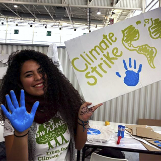 A young dark-skinned woman with long hair is smiling and looking at the camera. She is holding a poster with the words "Climate Strike." The palm of her right hand is painted blue, and her handprint is on the poster. She is sitting on a chair next to a table with poster boards, papers and blue paint.
