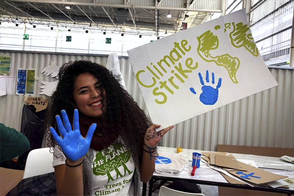 A young dark-skinned woman with long hair is smiling and looking at the camera. She is holding a poster with the words "Climate Strike." The palm of her right hand is painted blue, and her handprint is on the poster. She is sitting on a chair next to a table with poster boards, papers and blue paint.